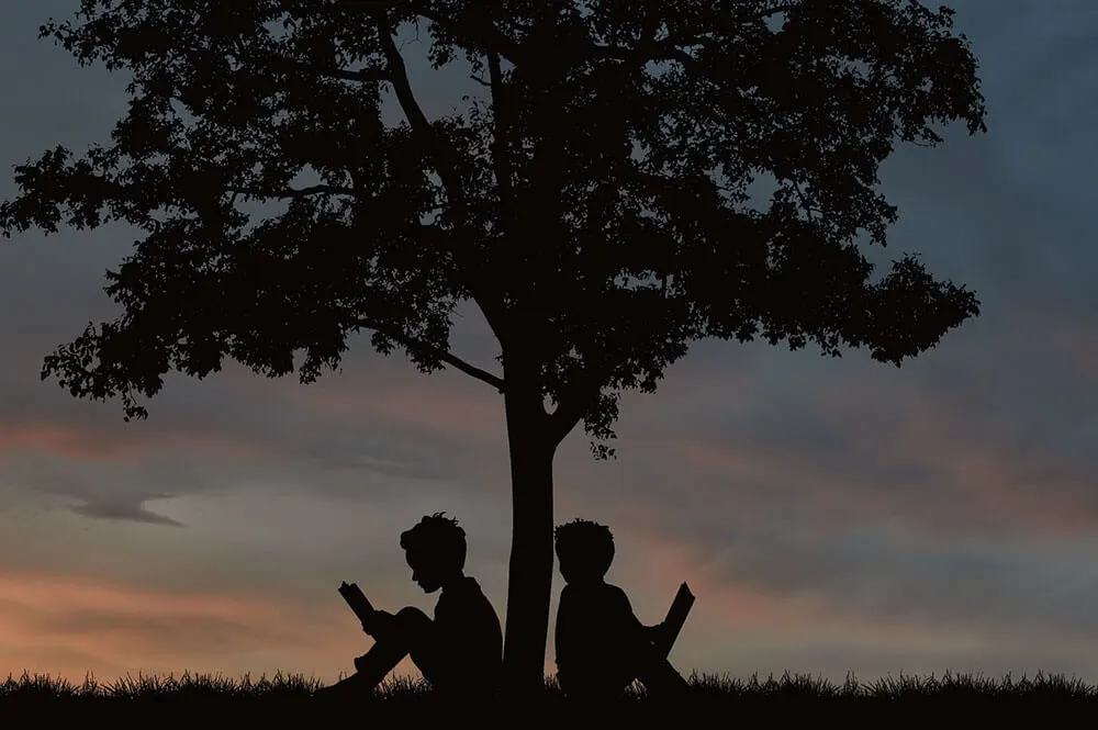 Silhouette di due bambini che leggono sotto un grande albero al tramonto